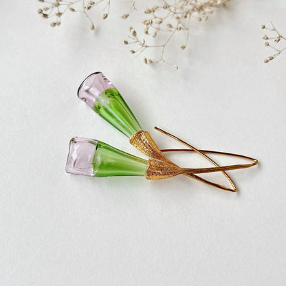 Pair of green and pale pink glass earrings with gold colored findings on grey background laying on side. Small dried flowers above image.
