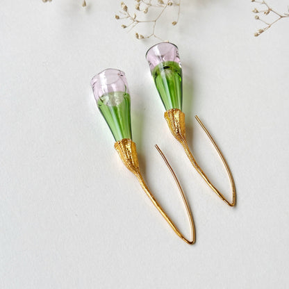 Pair of green and pale pink glass earrings with gold colored findings upside down, on grey background. Small dried flowers above image.