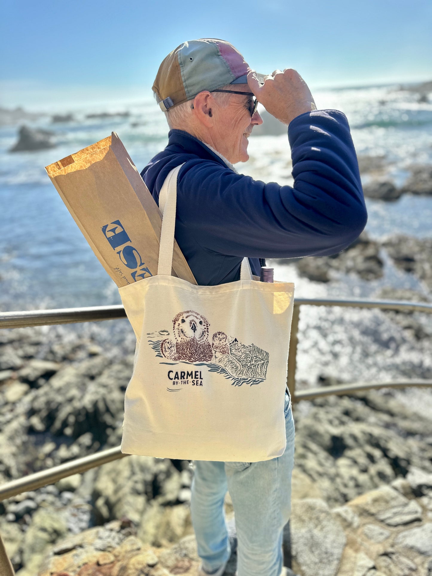 Man with hat, sunglasses and canvas tote bag with picture of otter pup wrapped in seaweed, and carmel-by-the-sea written below, looking in the distance.