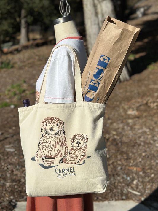Canvas tote bag with picture of a momma otter and pup, and carmel-by-the-sea written below on the shoulder of a manikin.