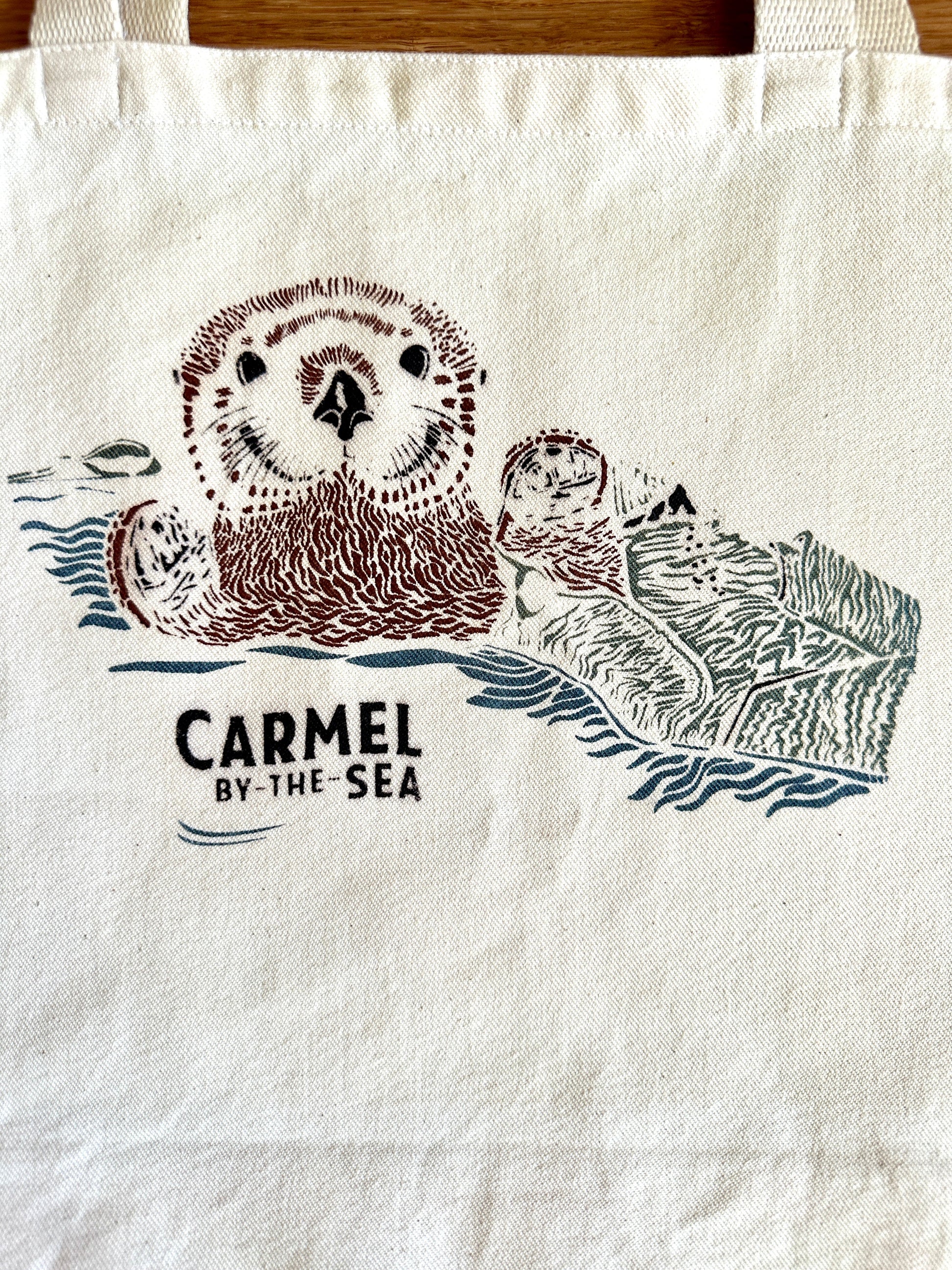 Closeup image of otter pup wrapped in seaweed, and carmel-by-the-sea written below.