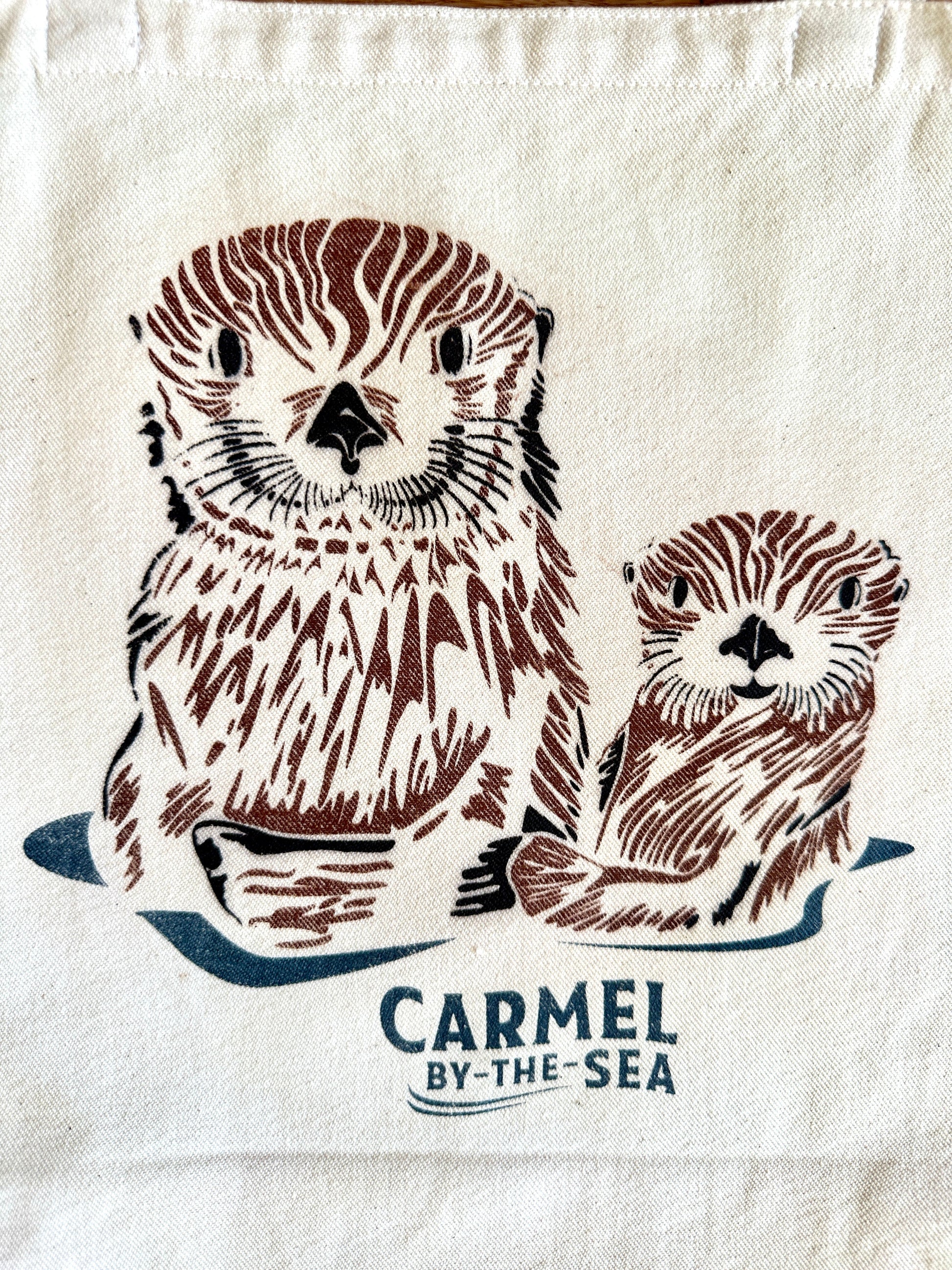 Closeup image of a momma otter and pup, and carmel-by-the-sea written below.
