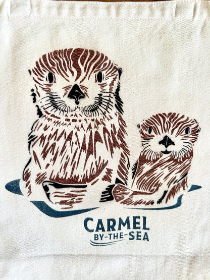 Closeup image of a momma otter and pup, and carmel-by-the-sea written below.