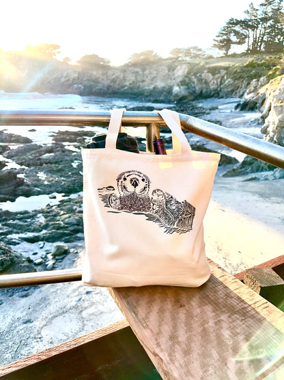 Canvas tote bag hanging on a rail, with picture of otter pup wrapped in seaweed.