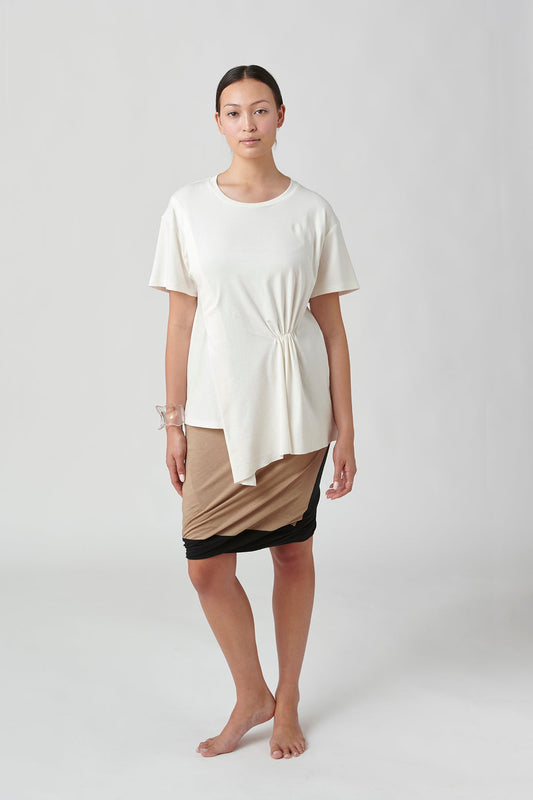 Front view of woman wearing white t-shirt, left side slightly longer than left side.  Also wearing tan skirt with black trim.