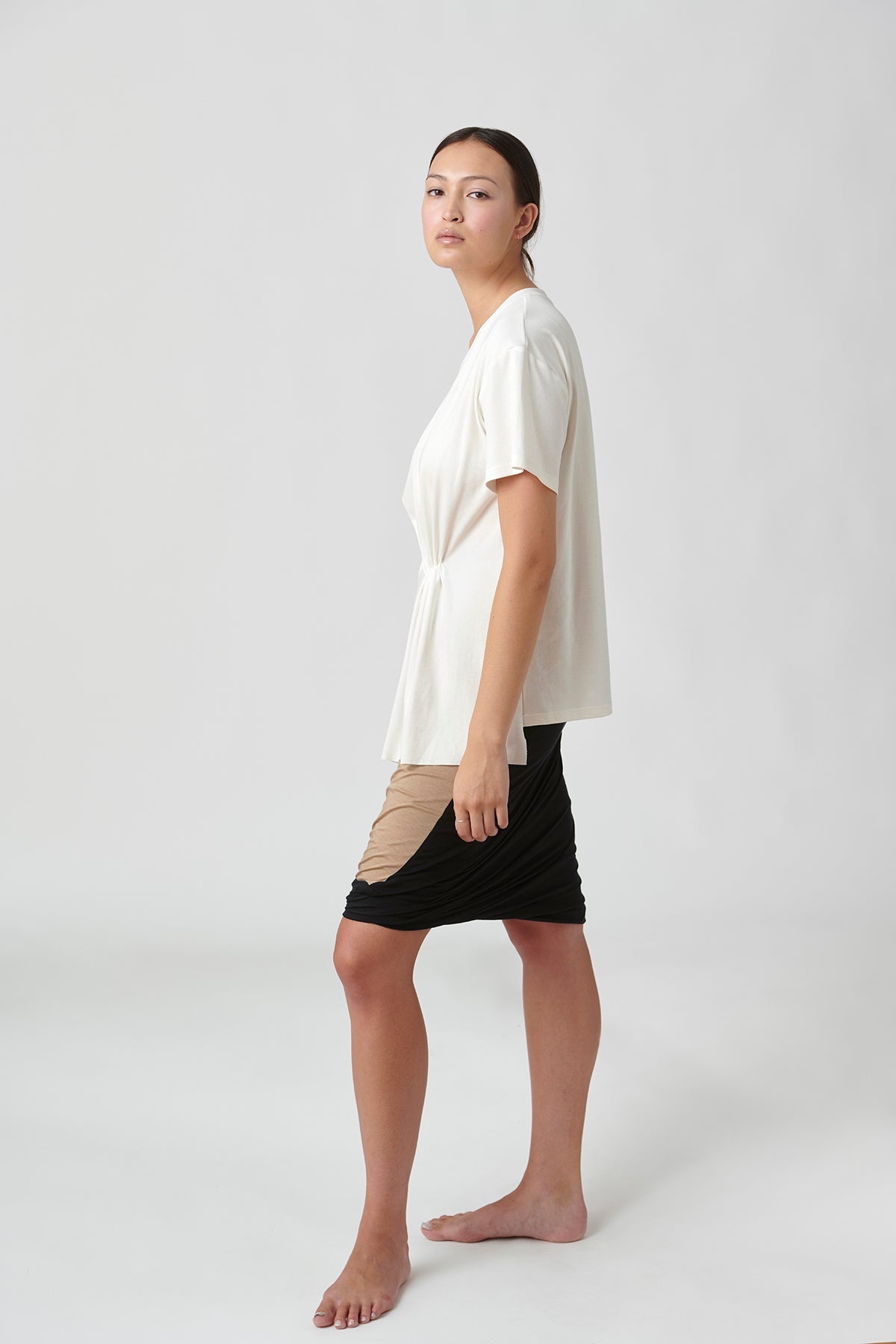 Front left view of woman wearing white t-shirt, left side slightly longer than left side.  Also wearing tan skirt with black trim.