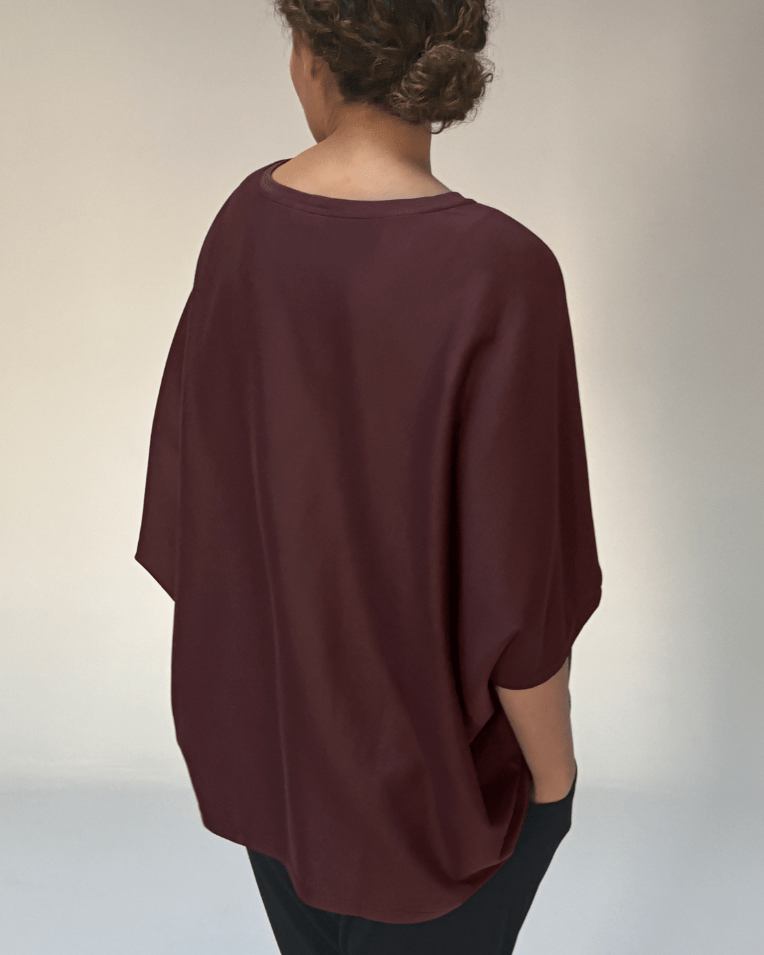 A woman shown from the back wearing a loose, draped short-sleeve top in a deep burgundy or maroon shade, paired with black pants, against a plain light gray studio background.