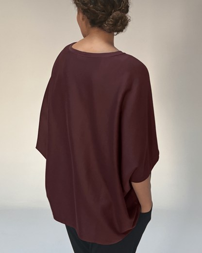 A woman shown from the back wearing a loose, draped short-sleeve top in a deep burgundy or maroon shade, paired with black pants, against a plain light gray studio background.