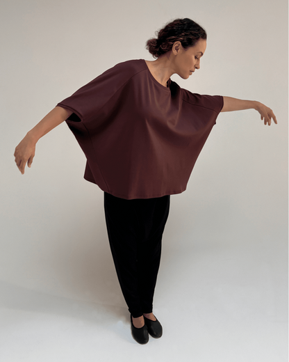 A woman modeling a loose, draped short-sleeve top in deep burgundy, paired with black pants and black flats, posed against a neutral light gray studio background.