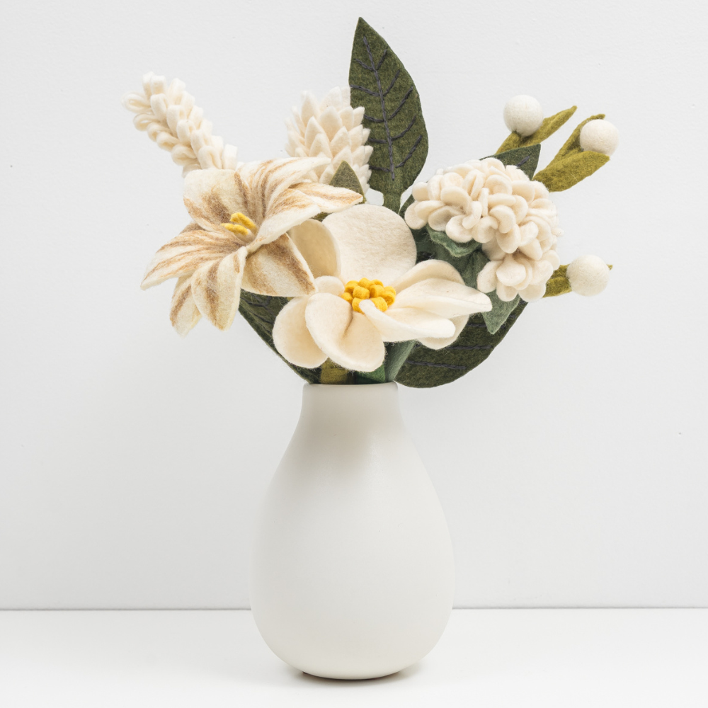 Bouquet of white felt flowers and green leaves in a white vase with white background.