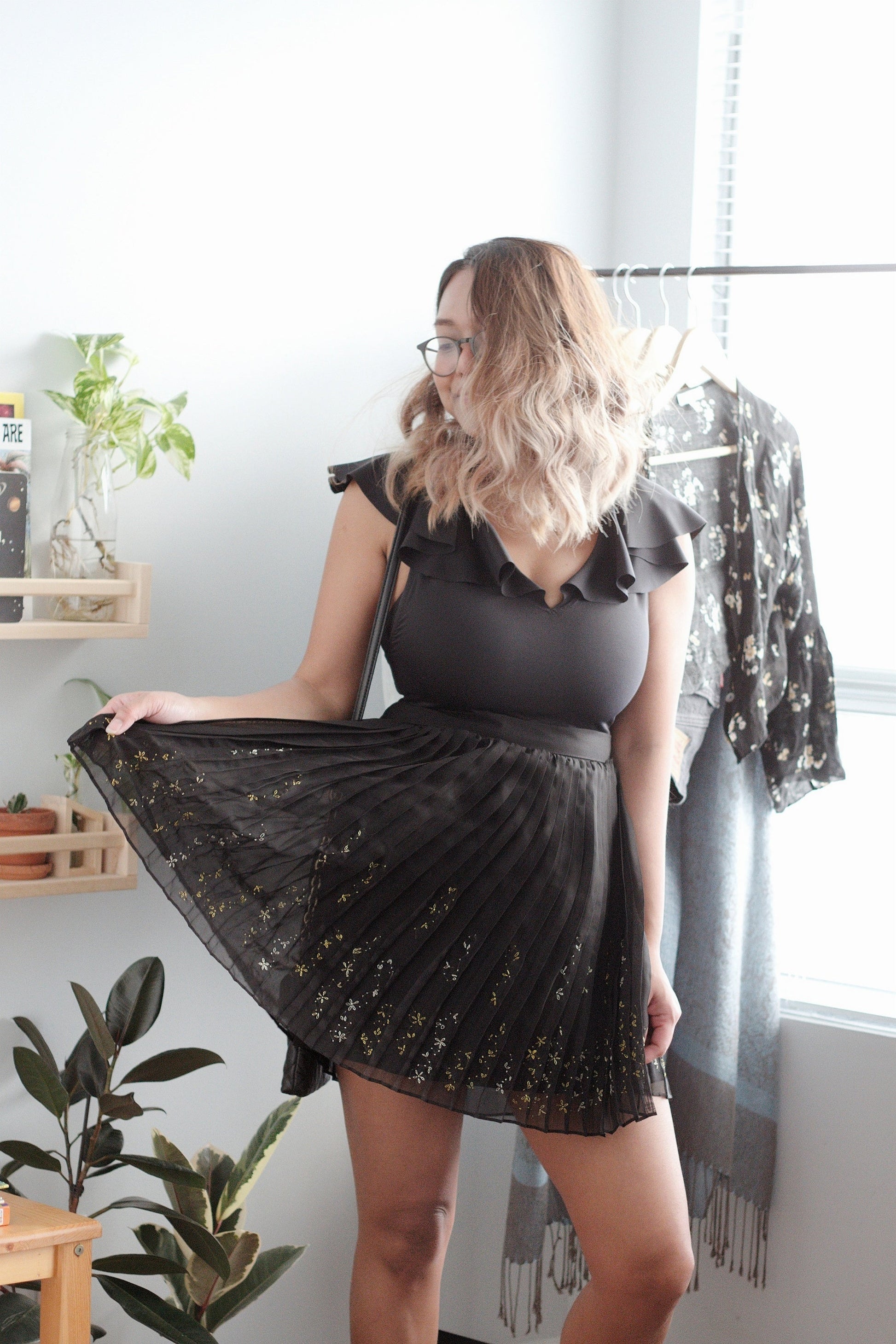 Woman wearing a black dress with a sheer overlay in a room with plants and a window.