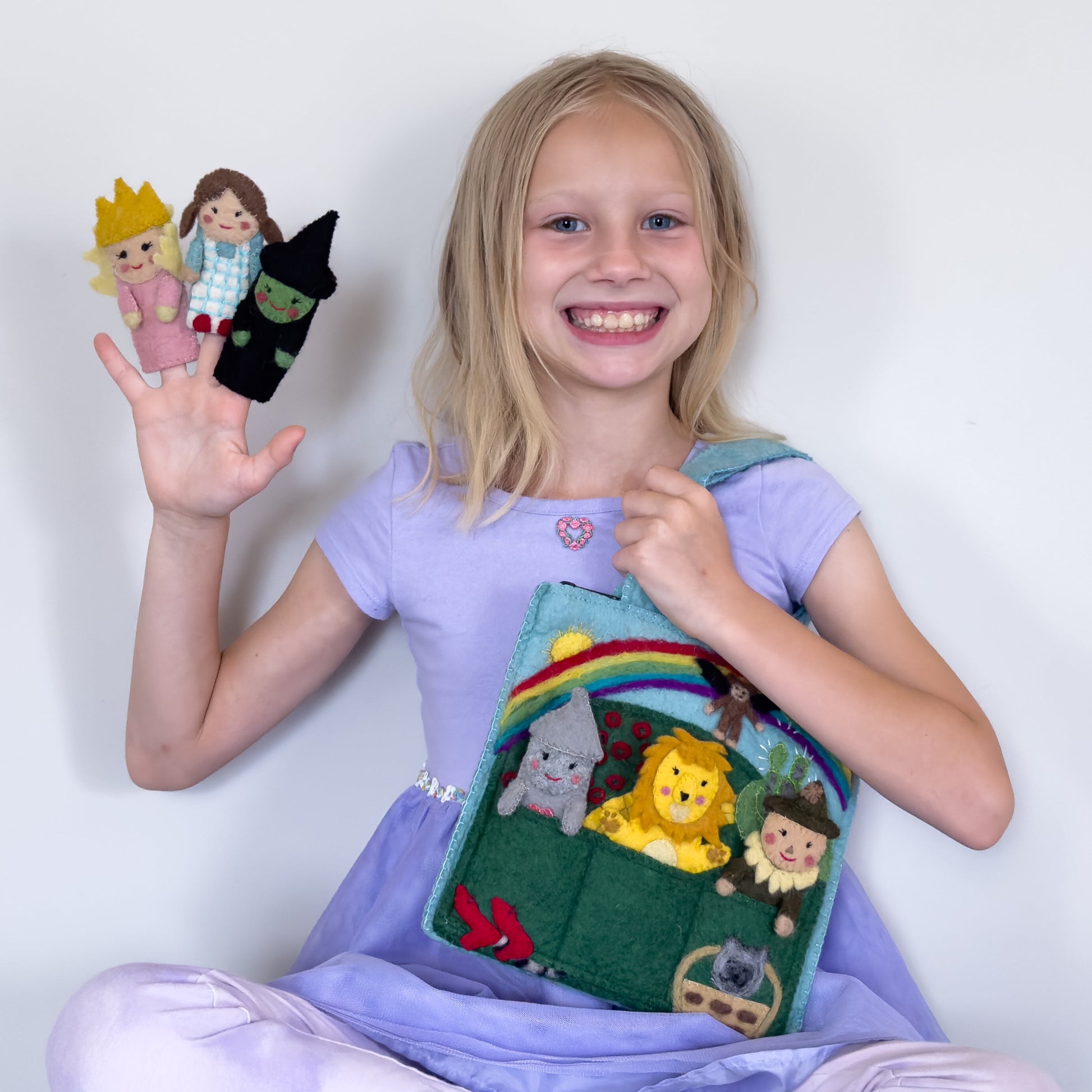 Young girl in purple dress holding a felt puppet bag for kids with wizard of oz themed characters of Dorothy, the Tin Man, The Cowardly Lion, Glinda the Good Witch, The Wicked Witch of the West, and the Scarecrow.