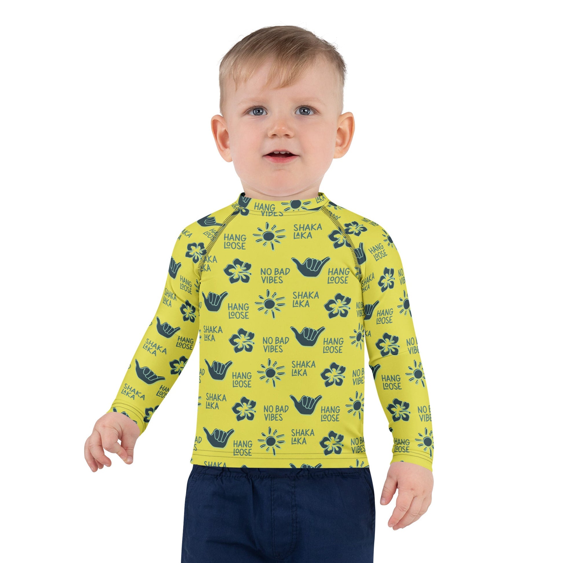 Toddler boy wearing a yellow rash guard with navy designs of the sun, hibiscus flowers, hang loose hand sign, and the words hang loose, no bad vibes, and shaka laka.