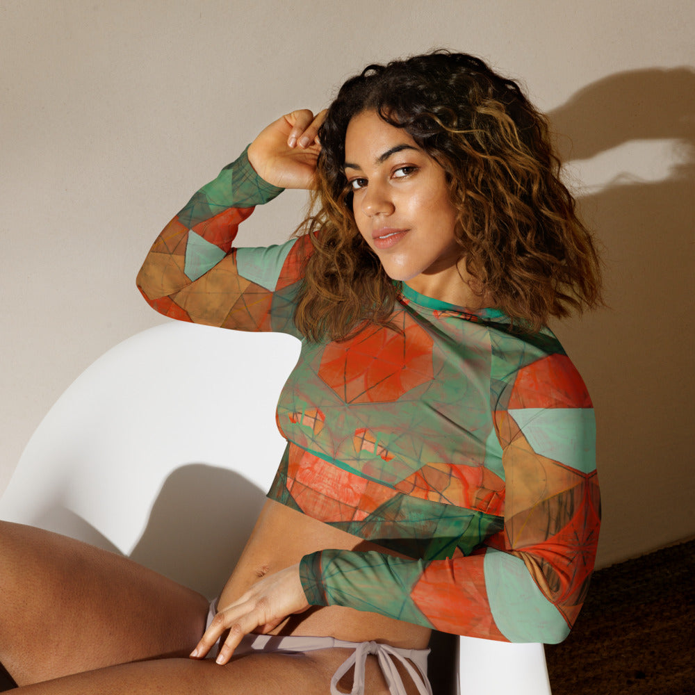 Woman sitting with hand in hair in a green and orange geometric print cropped rash guard and tan bikini bottoms.