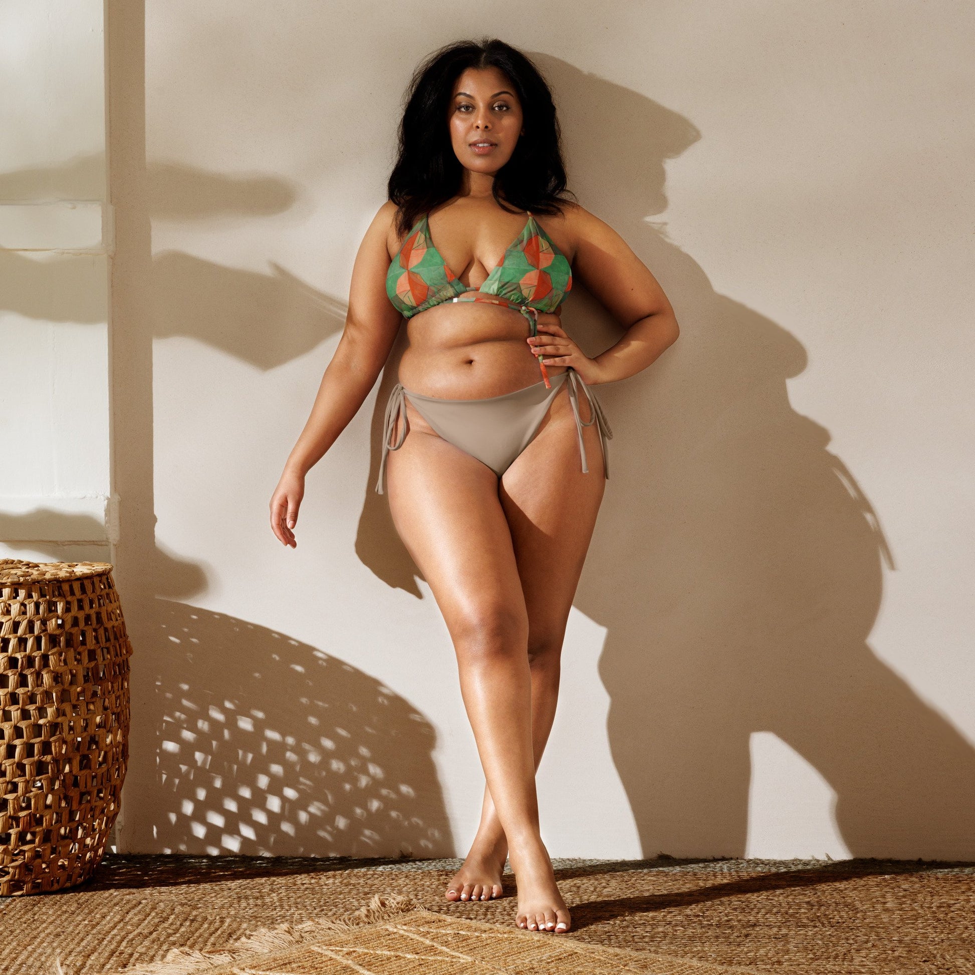 Woman leaning against shadowy wall, wearing light and dark green colored print bikini top with orange flowers and light mocha bottoms.