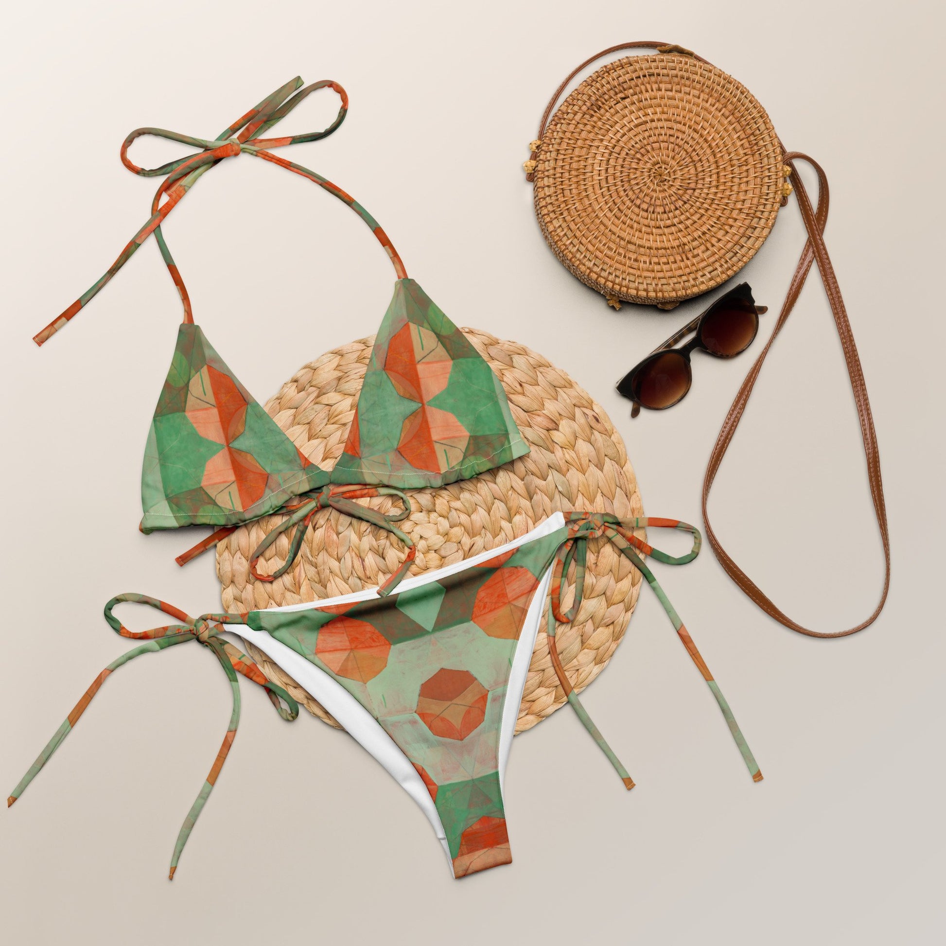 Light and dark green colored print bikini with orange geometrical flowers laying on a straw mat, next to a straw purse and sunglasses.