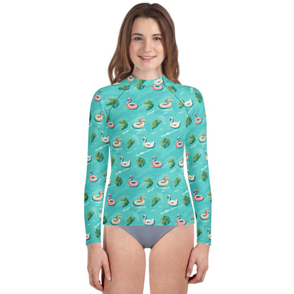 Girl wearing an aqua blue rash guard with images of pink floaty toy unicorns, swans, and Flamingos and green tropical leaves.