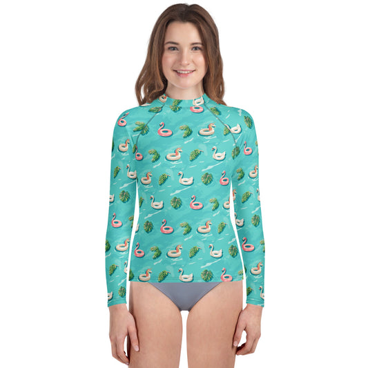 Girl wearing an aqua blue rash guard with images of pink floaty toy unicorns, swans, and Flamingos and green tropical leaves.