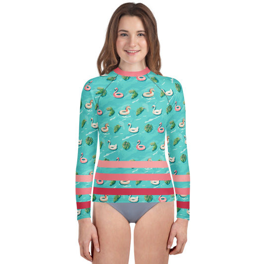 Girl wearing aqua blue rash guard with pink neckline and pink stripes on the lower half and images of pink floaty toy unicorns, swans, and Flamingos and green tropical leaves.