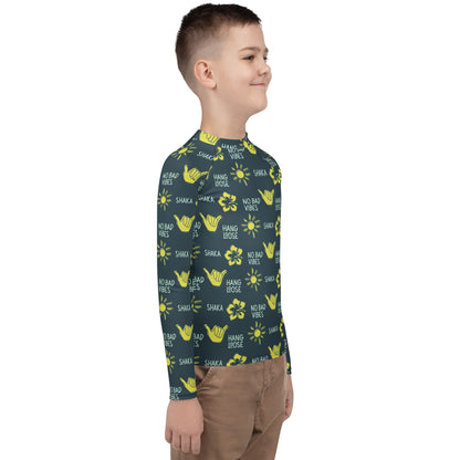 Side view of boy in dark blue rash guard with yellow designs of the sun, hibiscus flowers, hang loose hand sign, and the words hang loose, no bad vibes, and shaka.