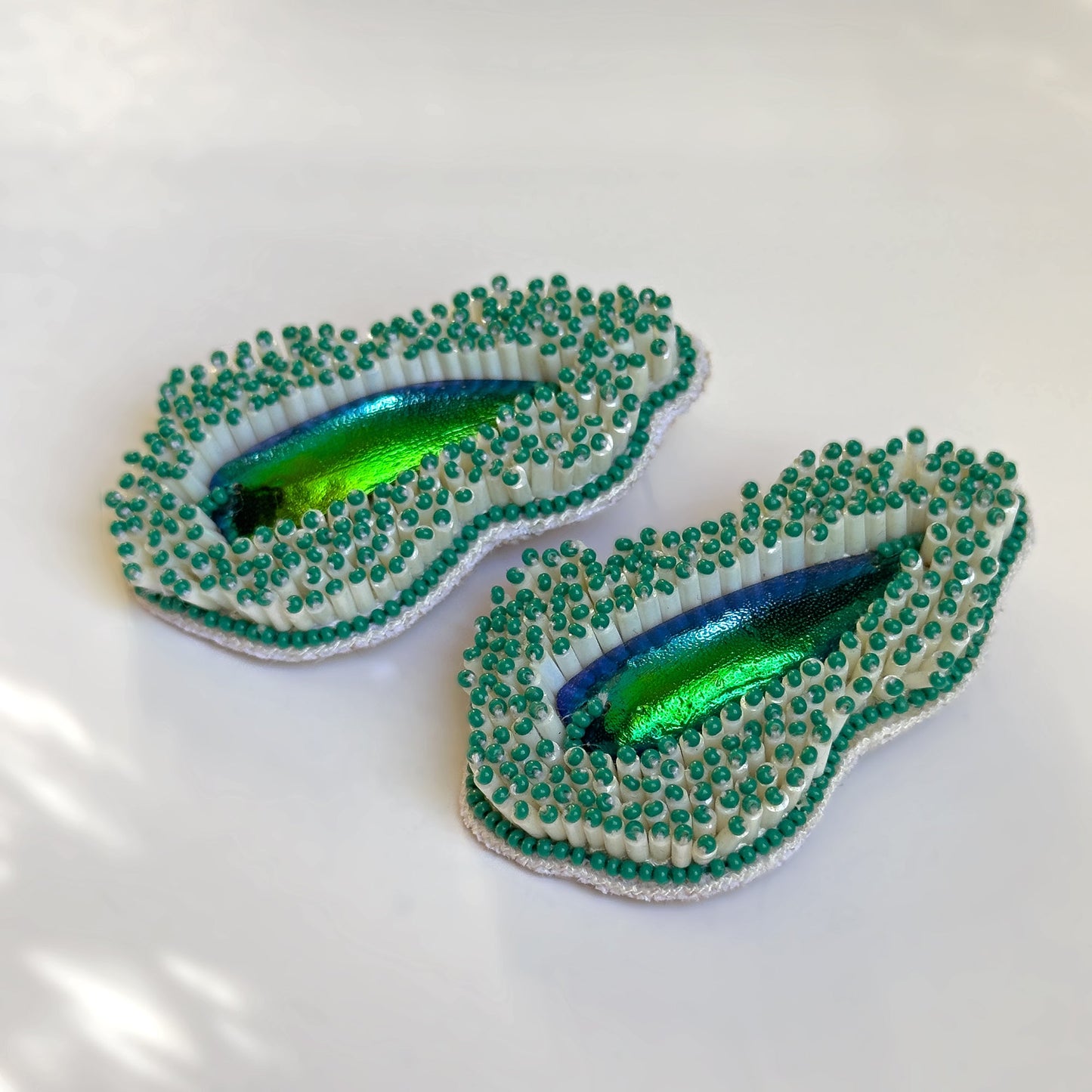 Pair of Pennata beaded green glass earrings with iridescent centers on shadowy background.