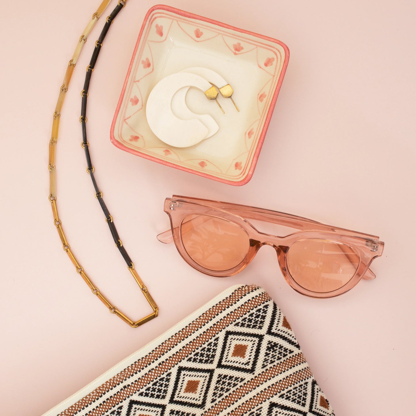 White, gold, and black link necklace next to a bowl with white hoop earrings, pink sunglasses, and a woven geometric cosmetic bag on a pink background.
