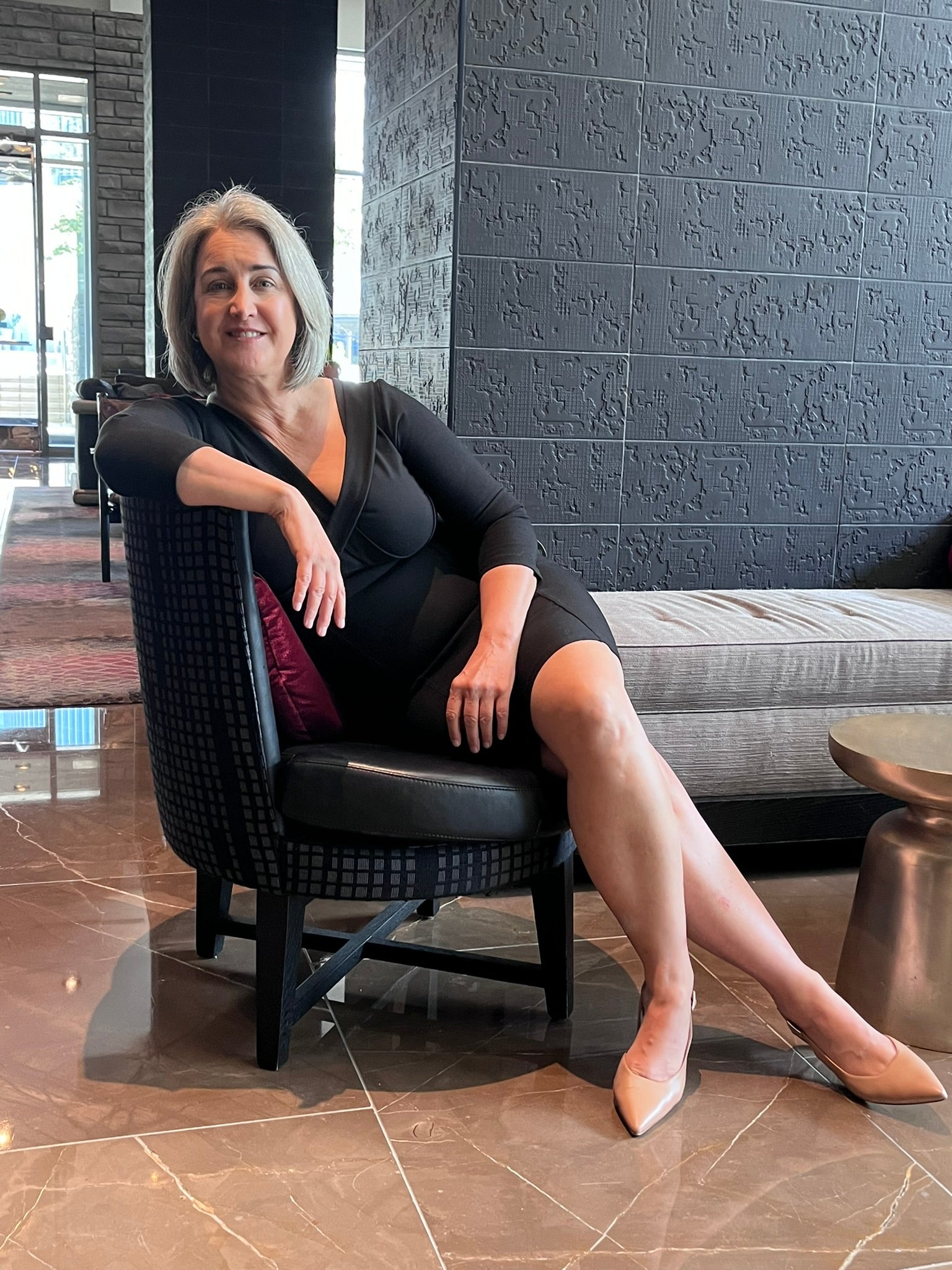 Relaxed woman sitting on black chair, leaning arm over back support, legs stretched and crossed, wearing black v neck dress.