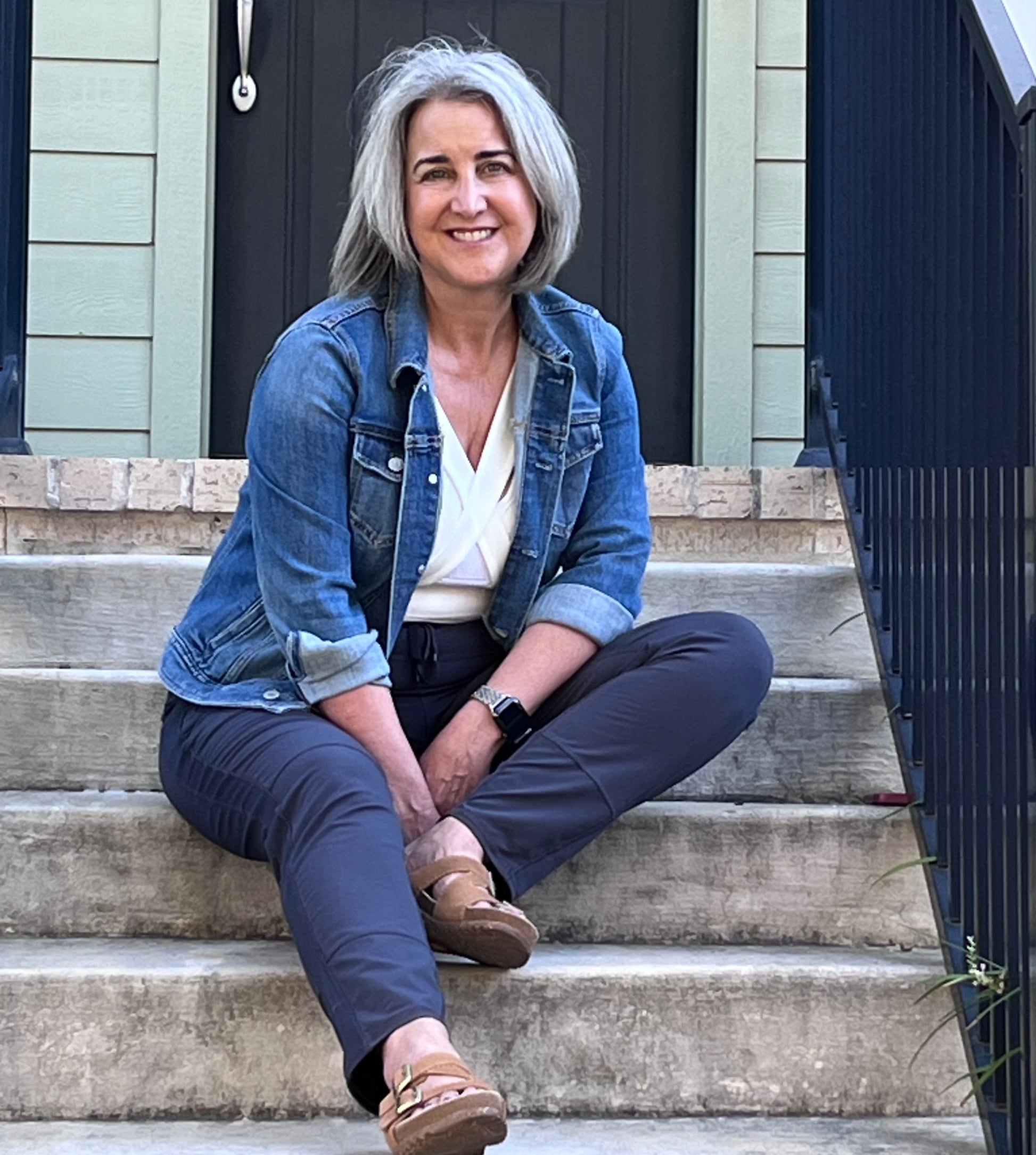 Woman sitting on outdoor steps, wearing white waist length crop top, jean jacket, and black pants.
