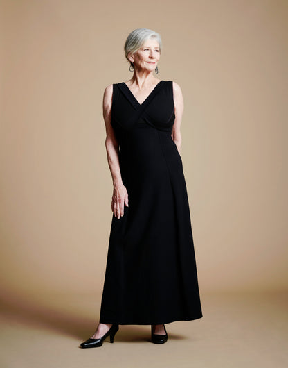 Woman wearing long black dress with lay-flat v neck look, right hand on thigh, left hand behind.