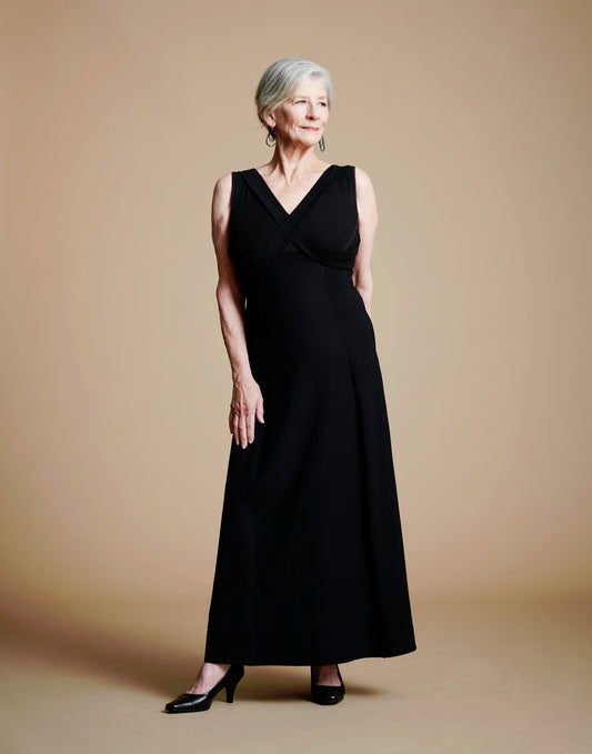 Woman wearing long black dress with lay-flat v neck look, right hand on thigh, left hand behind.