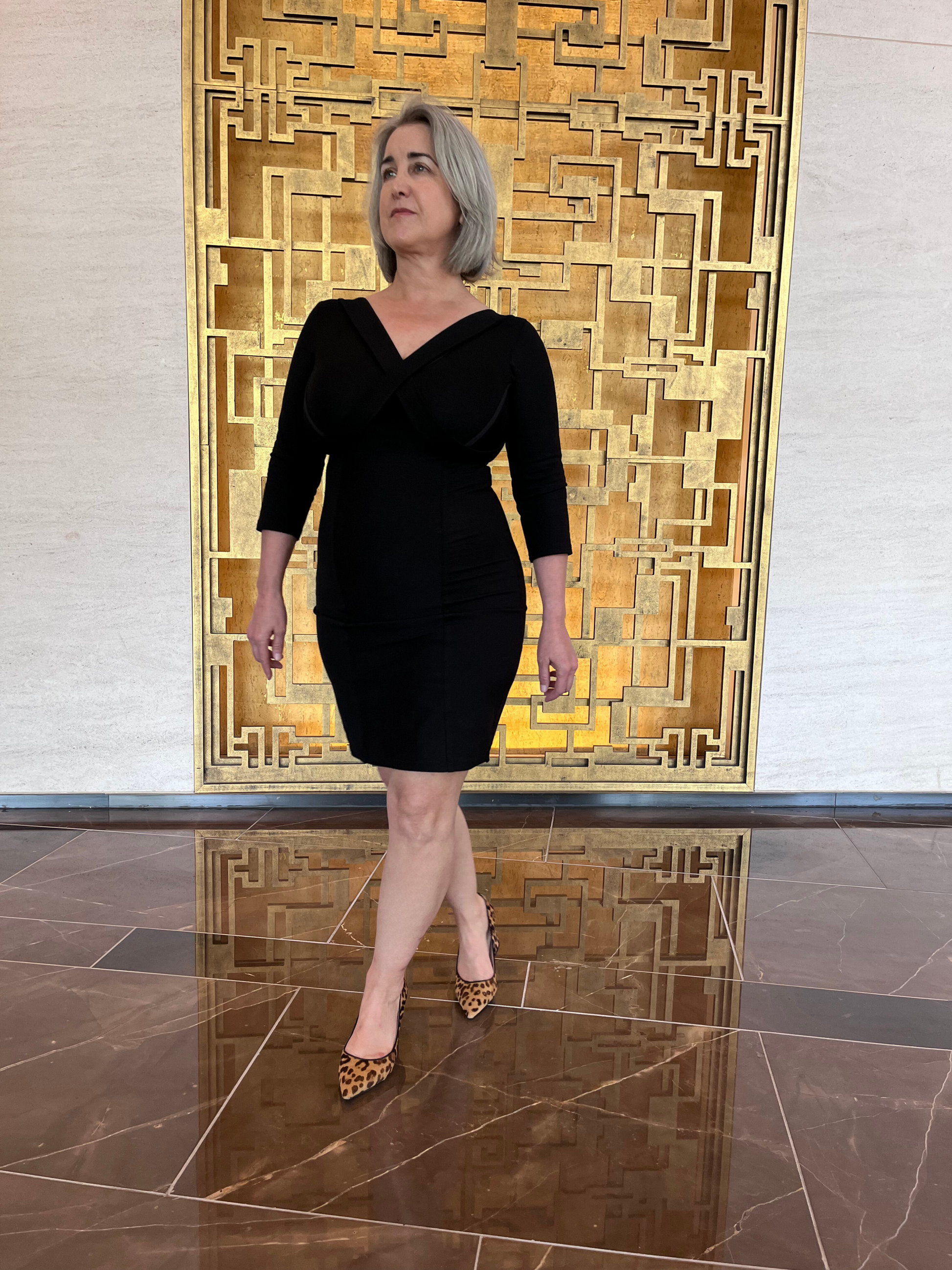Woman standing in front of gold grid, on brown marble floor, wearing black v neck dress.