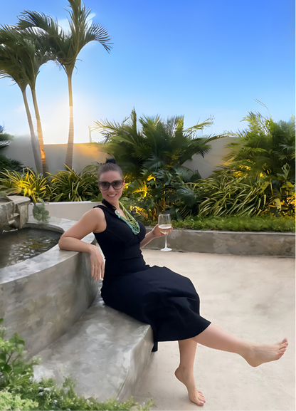 Woman sitting on stone fountain seat, glass in hand, wearing long black dress, palm trees in the background.
