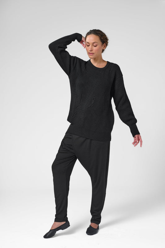 Woman with right elbow up wearing black crewneck sweater with asymmetric ribbed design and black pants.