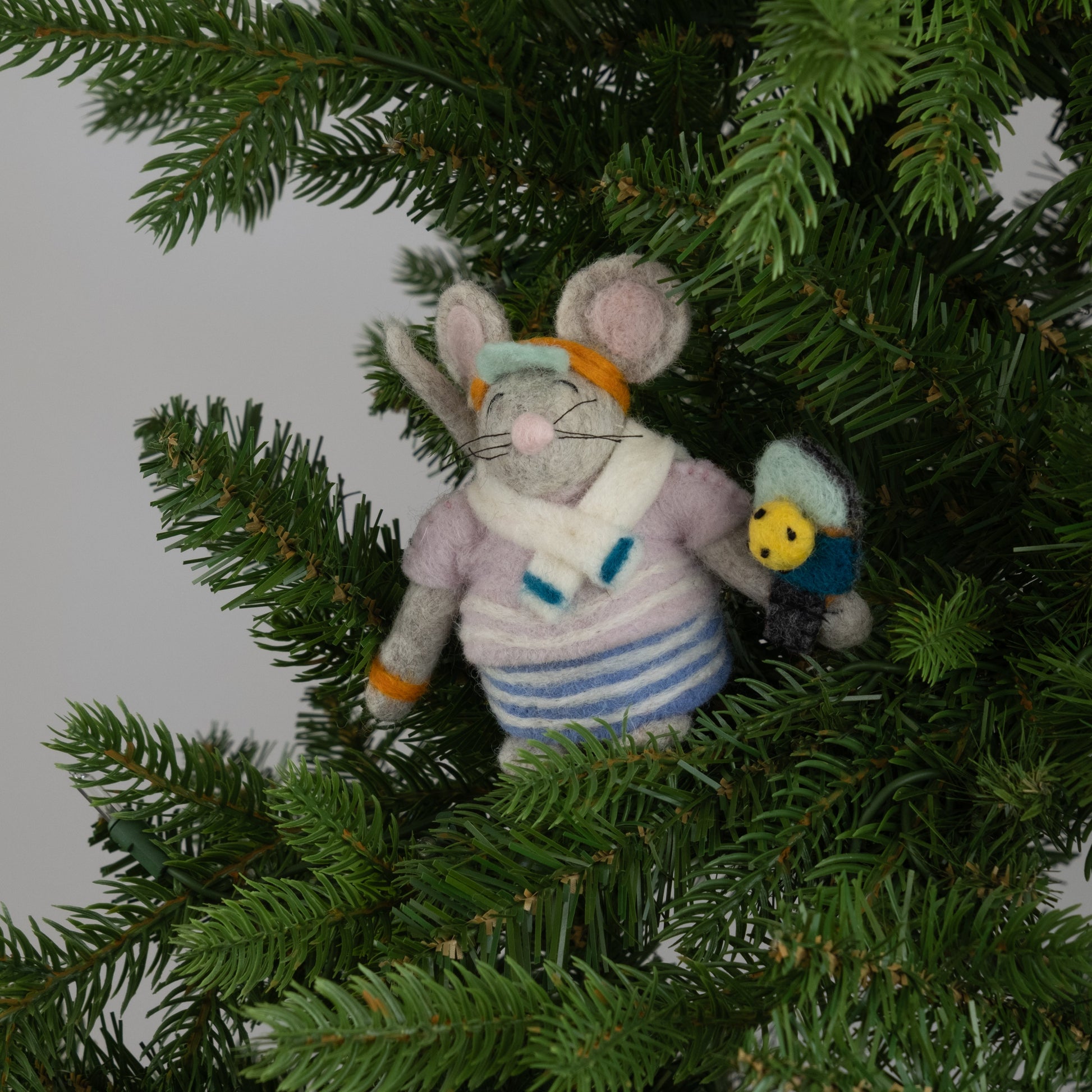 Felted ornament of a mouse dressed in a retro striped sweater, matching skirt, bright orange visor, and tiny sweatbands, with a pickleball paddle and ball in paw on a Christmas tree.