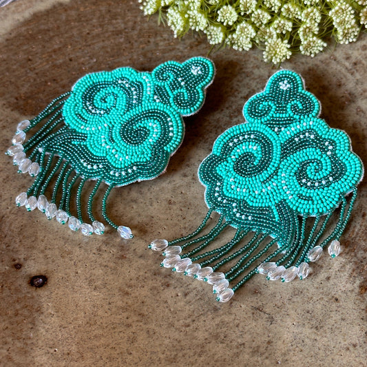 Pair of light and dark green cloud earrings with tassels on wood background.