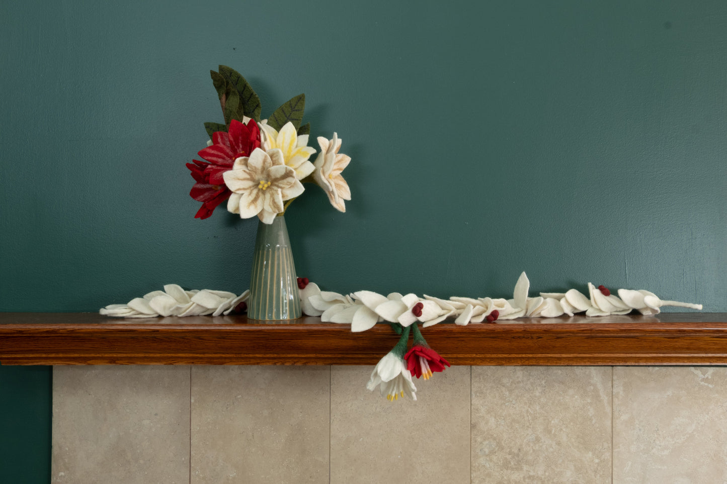 Felt ornaments of a red flower and a white flower hanging from a mantel decorated with Christmas flowers and garlands.