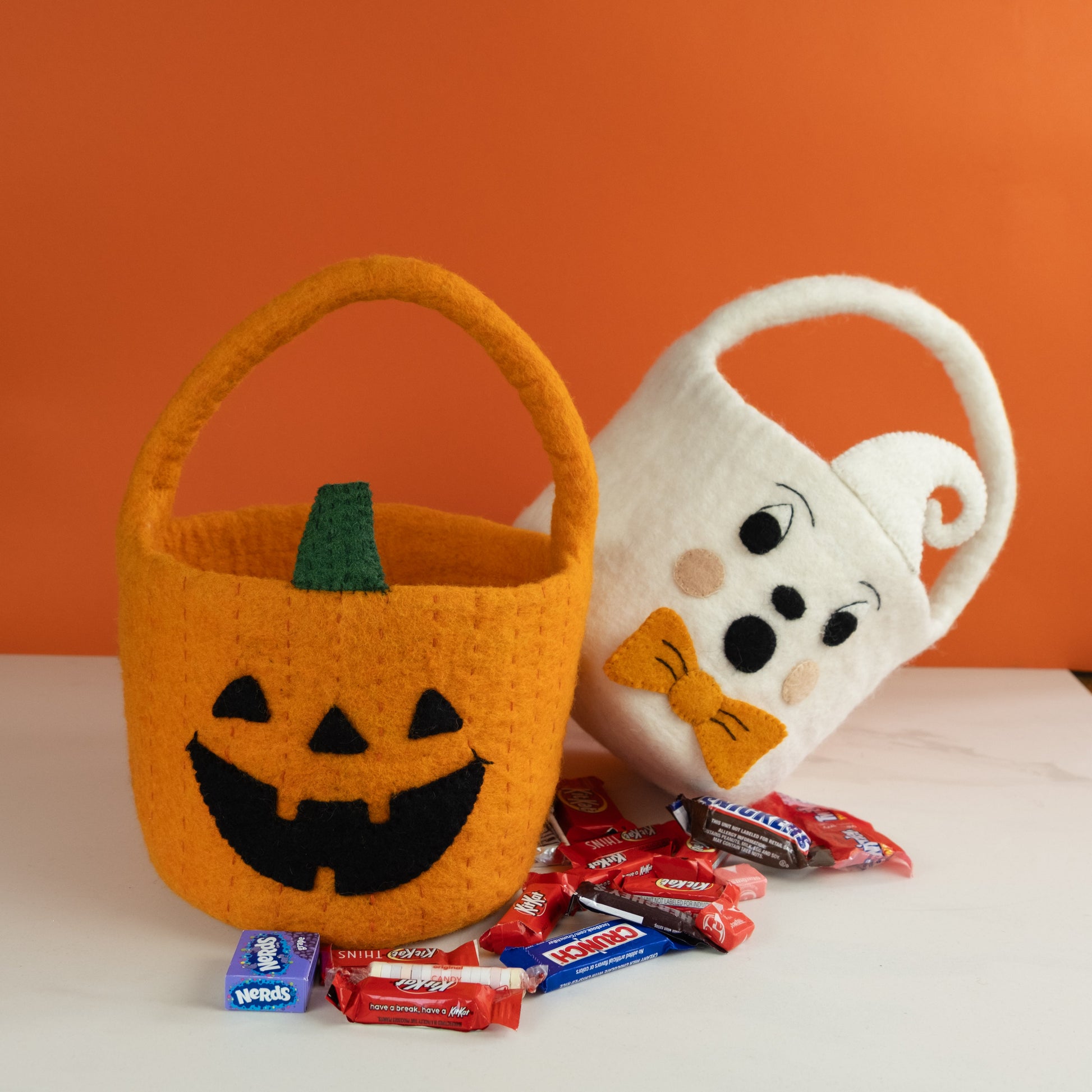 Orange felt jack-o-lantern trick-or-treat bucket and a white felt ghost trick-or-treat bucket with orange bow tie and candy below them.