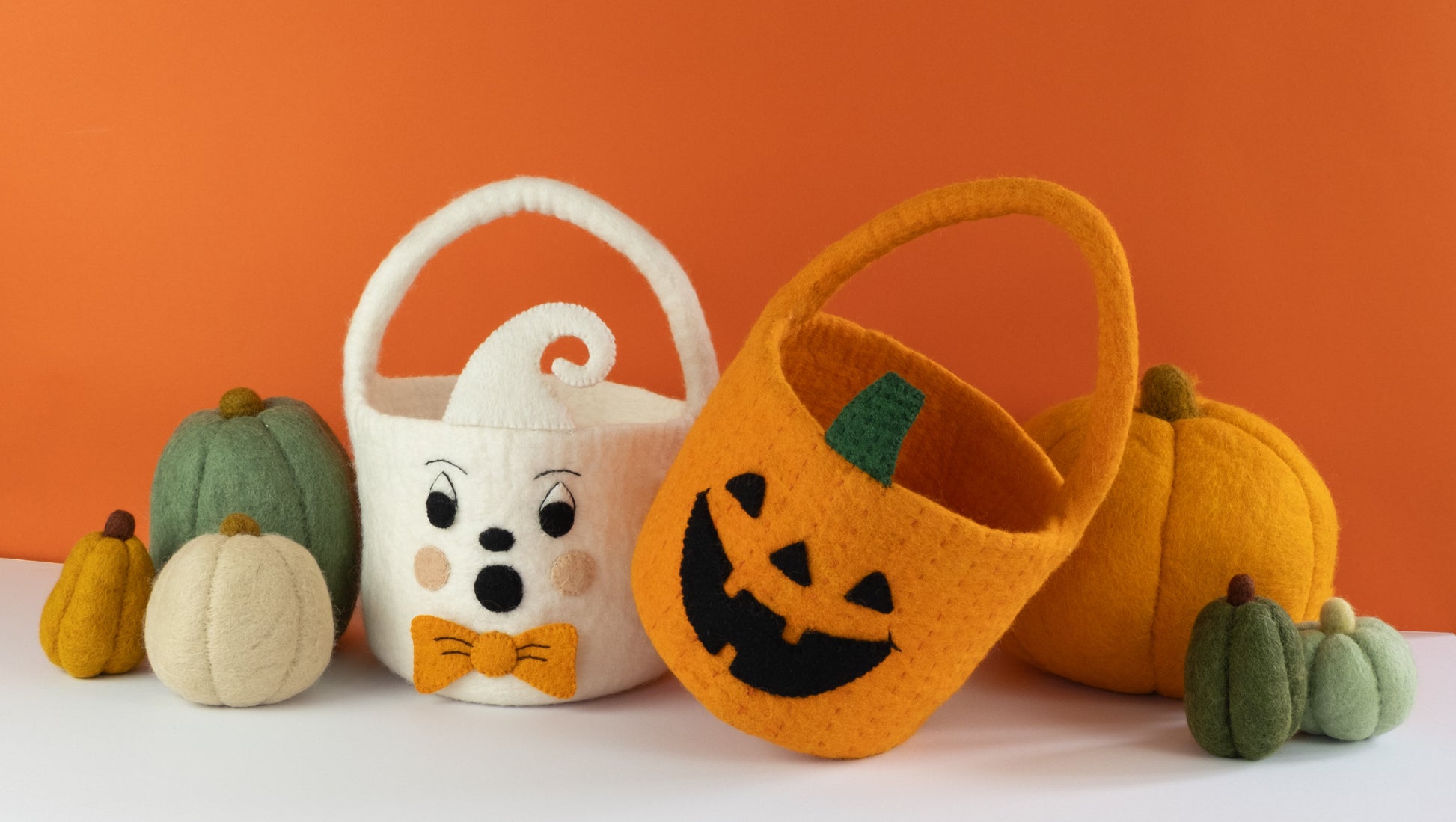 Orange felt jack-o-lantern trick-or-treat bucket and a white felt ghost trick-or-treat bucket with orange bow tie and felt pumpkins behind them.