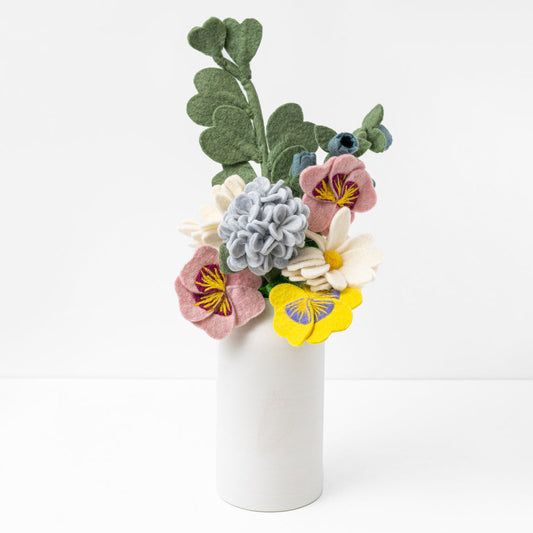 Felt flower bouquet of pink, blue, yellow, and white flowers with green leaves in white vase with white background.