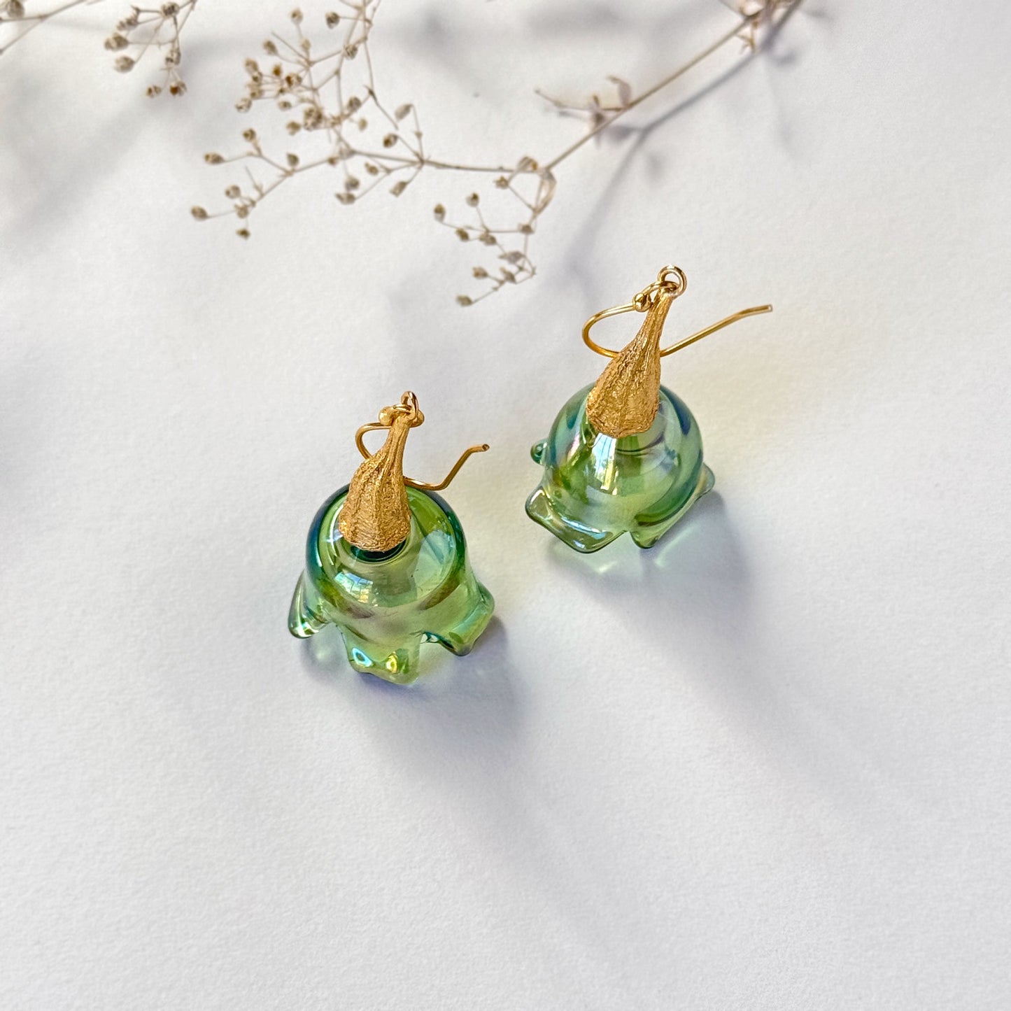 Pair of green pod glass earrings with gold findings standing upright on surface.  Small branch at top of image.