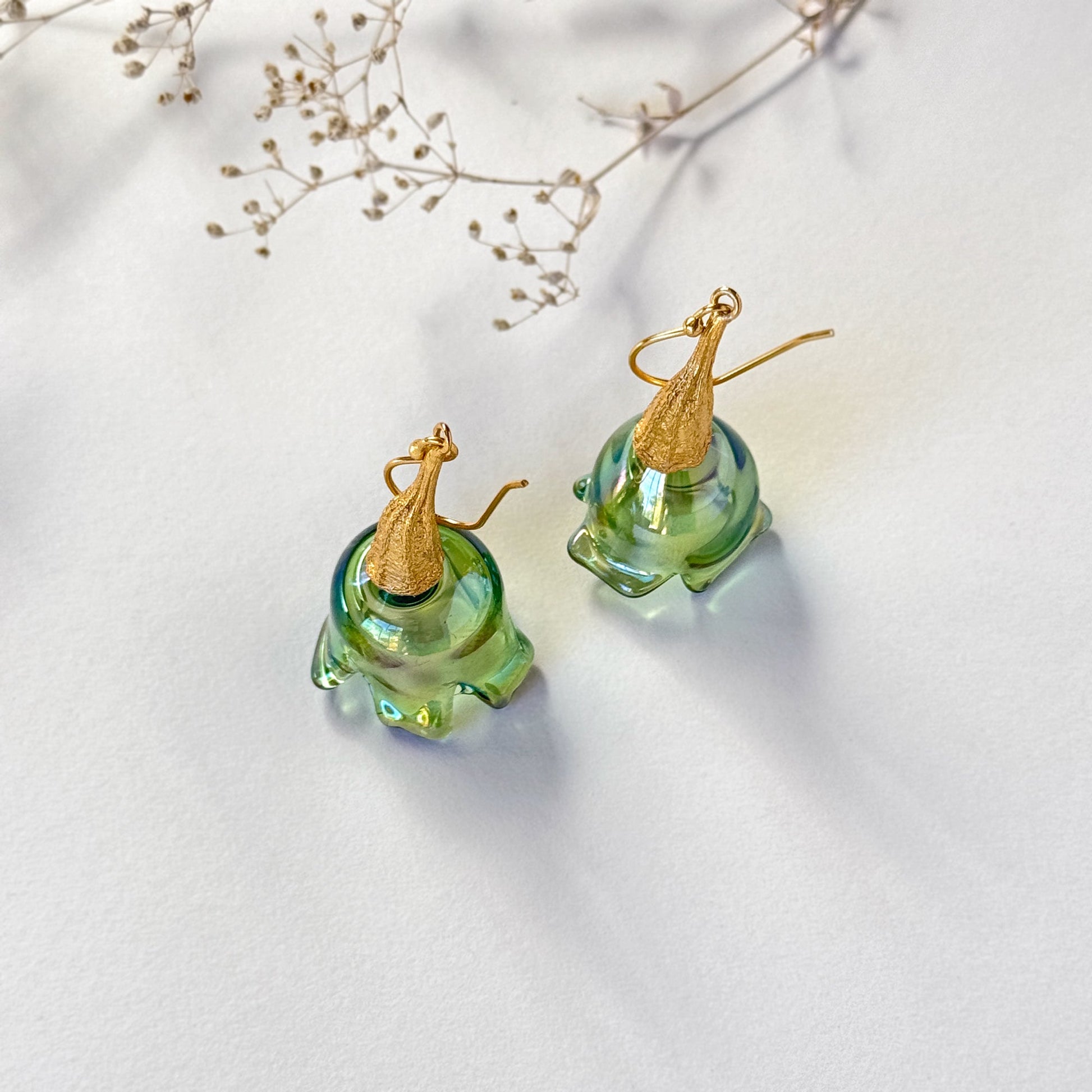 Pair of green pod glass earrings with gold findings standing upright on surface.  Small branch at top of image.