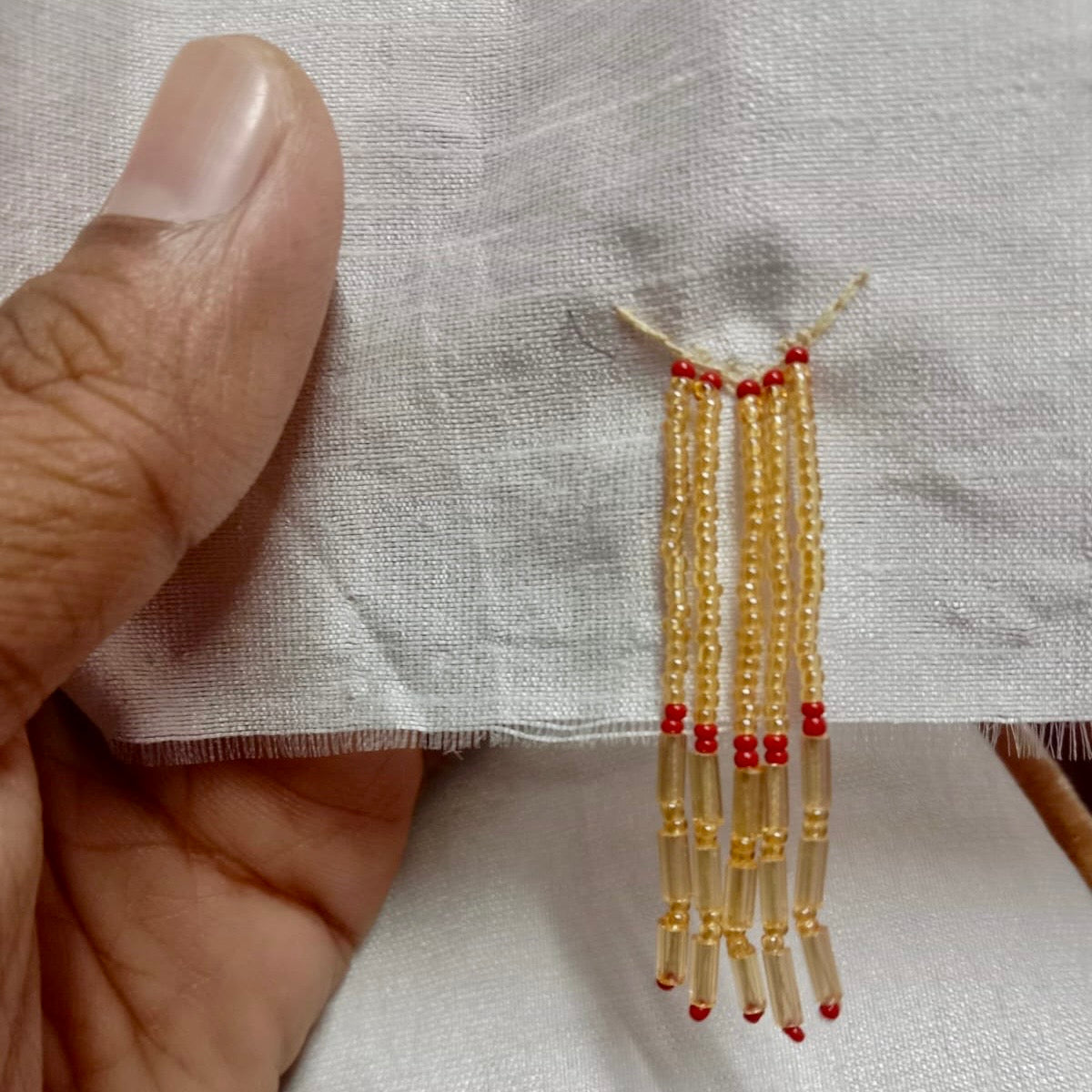 Left hand of person holding fabric while sewing white and gold glass bead earring with red accents.