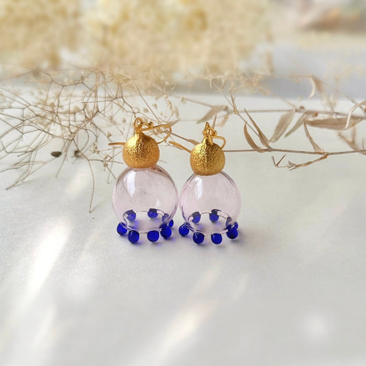 Pair of pale pink glass earrings with purple beads and gold plated findings on grey background.  Small dried flowers on upper section of image.