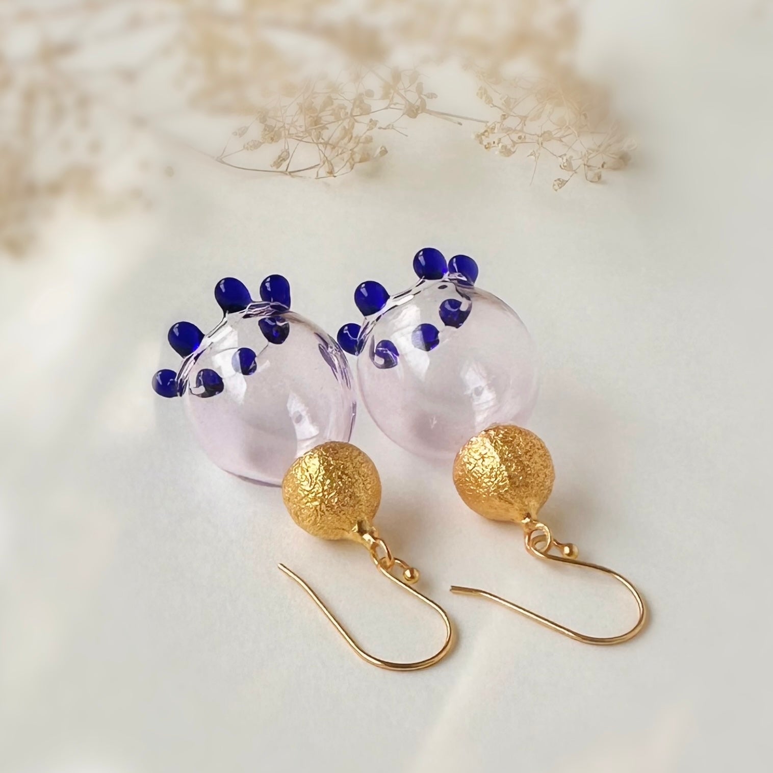 Pair of pale pink glass earrings laying upside down, with purple beads and gold plated findings. Small dried flowers in upper section of image.