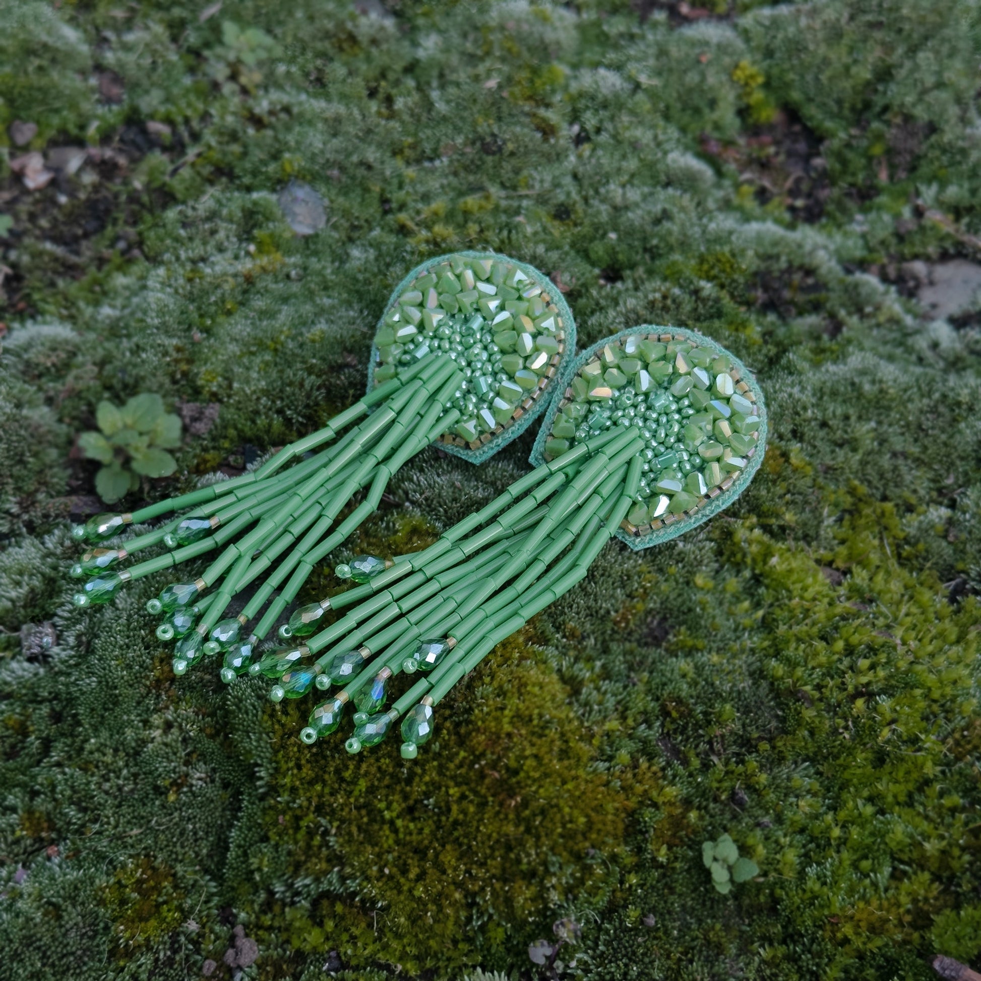 Pair of beaded green glass tassel earrings on moss background.