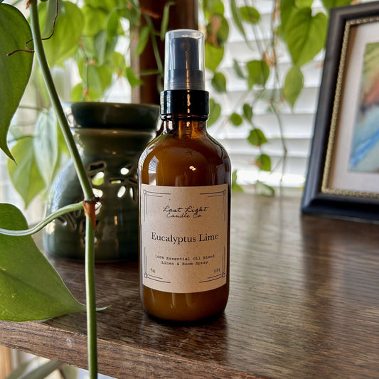New Izela Eucalyptus Lime Room Spray Primary Photo. Amber jar with spray top on wood table next to hanging plant.