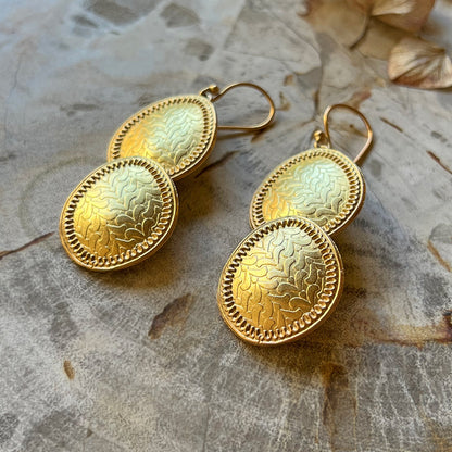 Pair of gold dangle earrings with etched leaf pattern on mottled background.