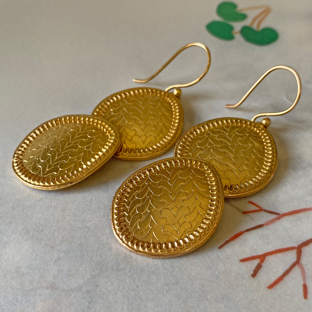 Pair of gold dangle earrings with etched leaf pattern on table.