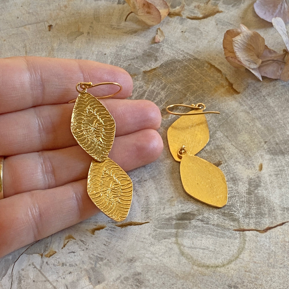 Left hand holding one of a pair of diamond shaped gold earrings.  Other earring on mottled surface.