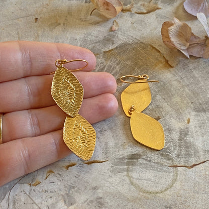 Left hand holding one of a pair of diamond shaped gold earrings.  Other earring on mottled surface.